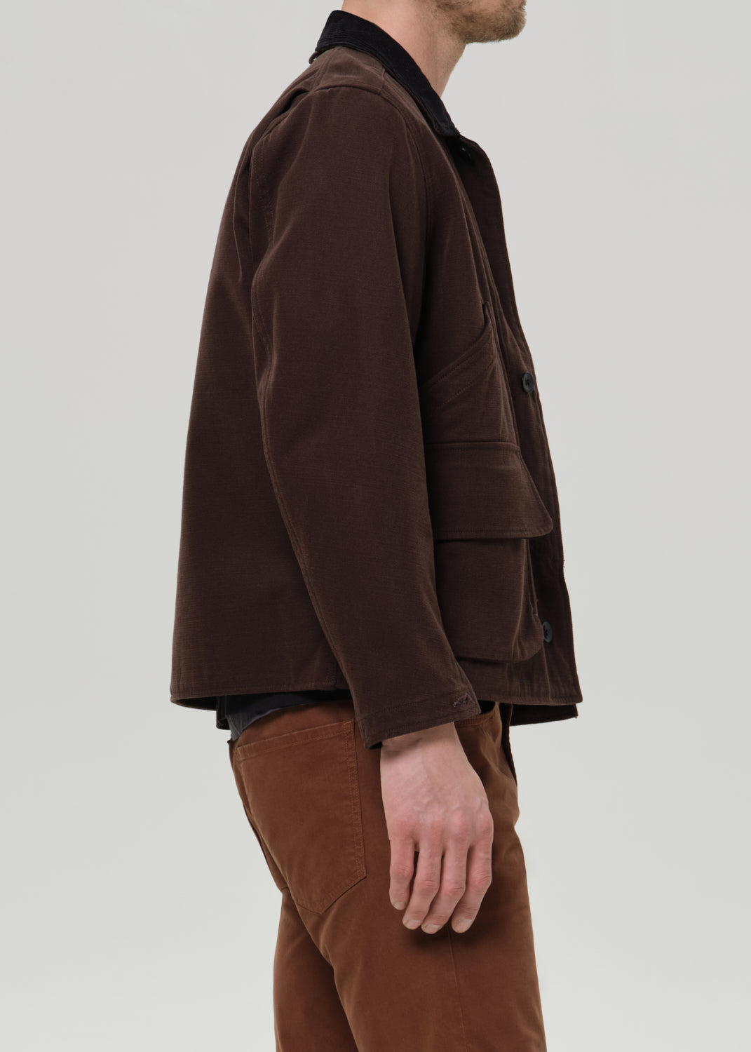 A man shown in side profile wearing a dark brown corduroy jacket over a black shirt and brown trousers, photographed against a grey background.