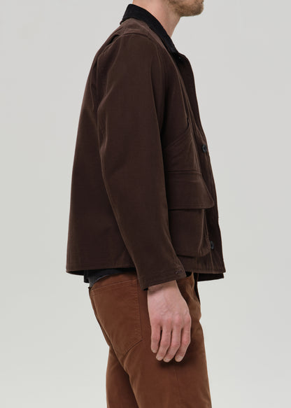 A man shown in side profile wearing a dark brown corduroy jacket over a black shirt and brown trousers, photographed against a grey background.