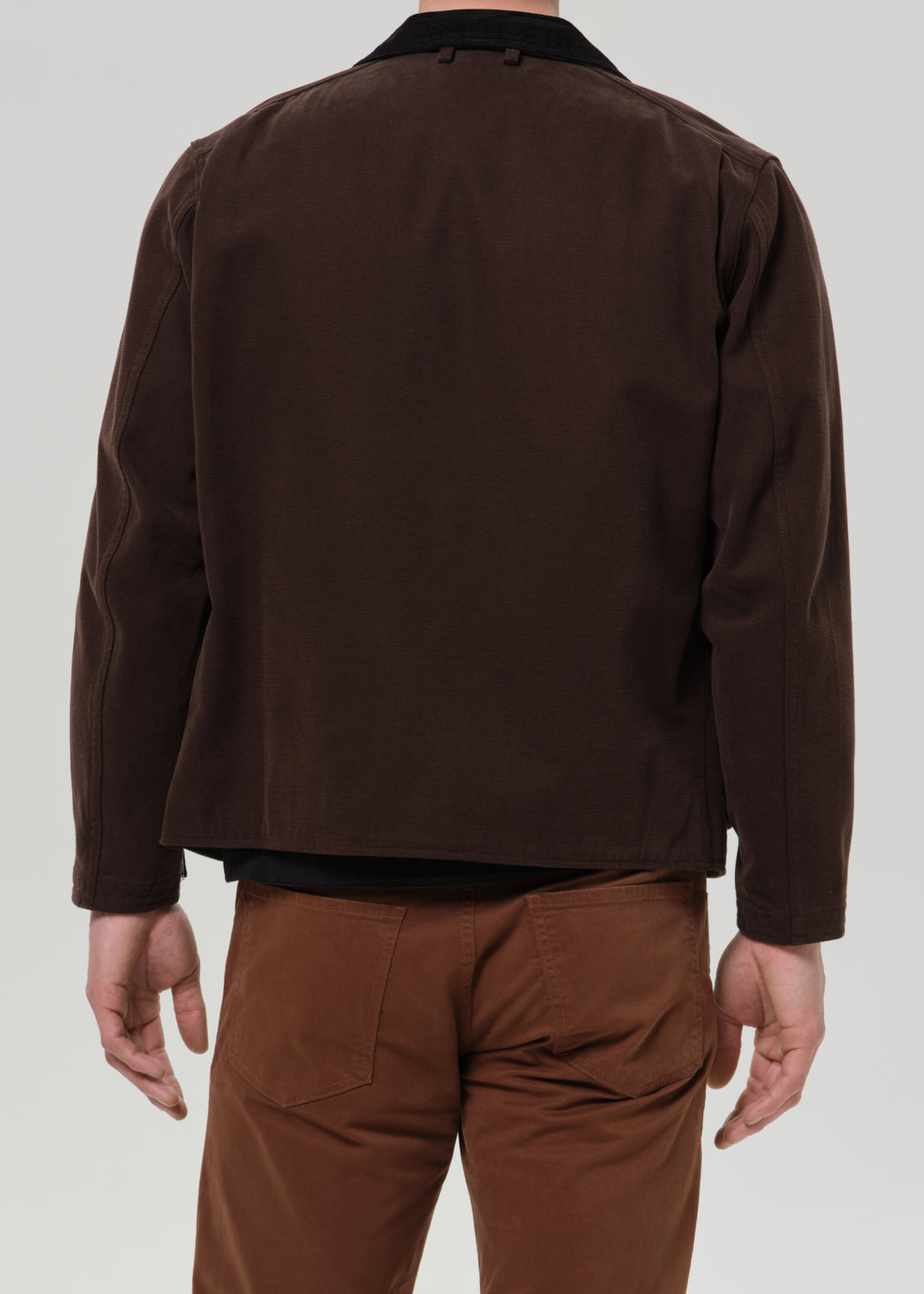 A man viewed from the back wearing a dark brown jacket with a black collar and brown pants, against a grey background.