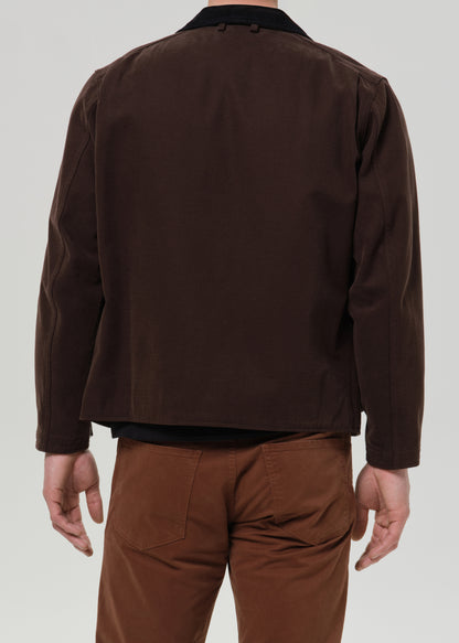 A man viewed from the back wearing a dark brown jacket with a black collar and brown pants, against a grey background.