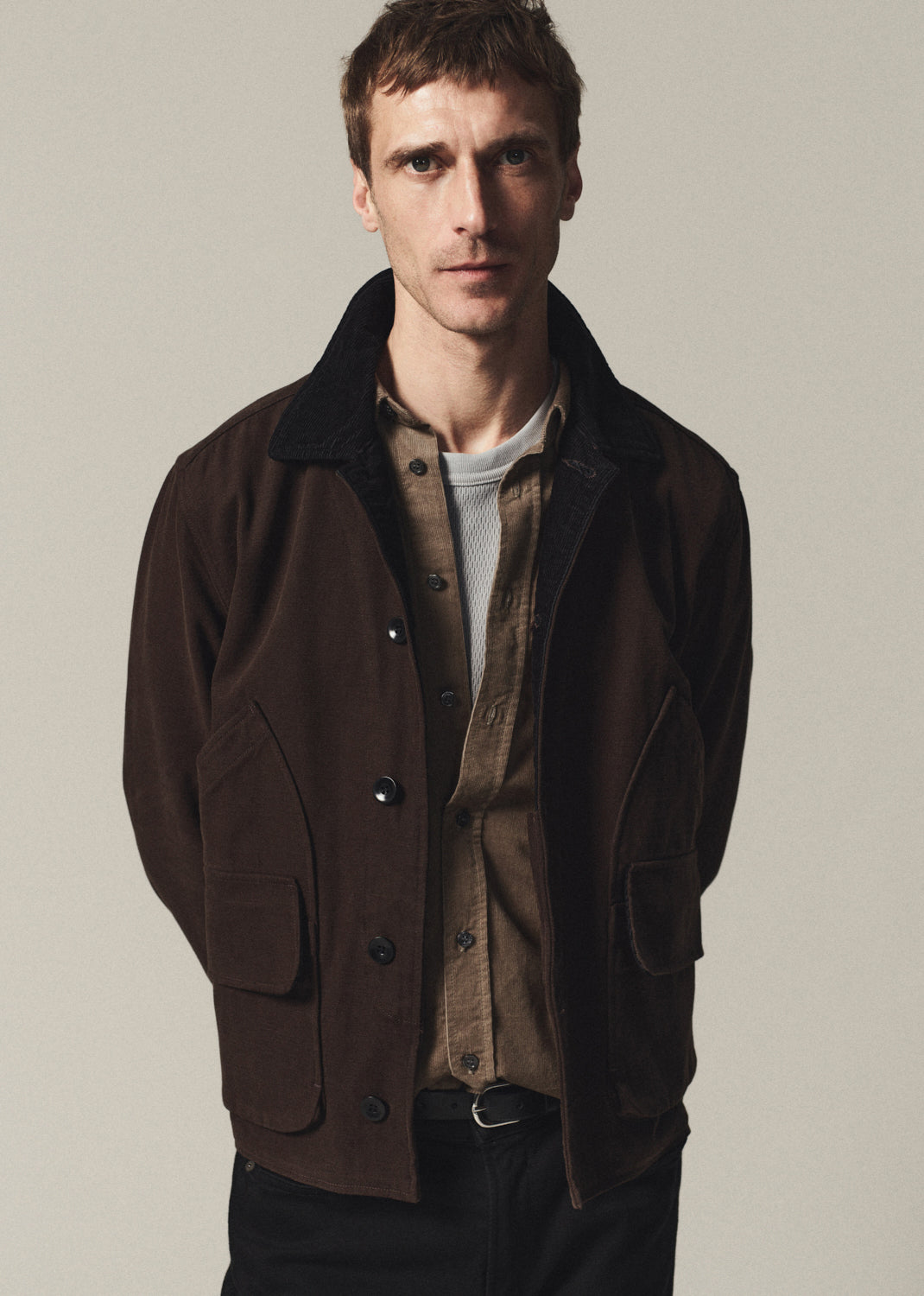A man wearing a dark brown corduroy jacket over a brown shirt and light gray t-shirt, standing against a plain light gray background.