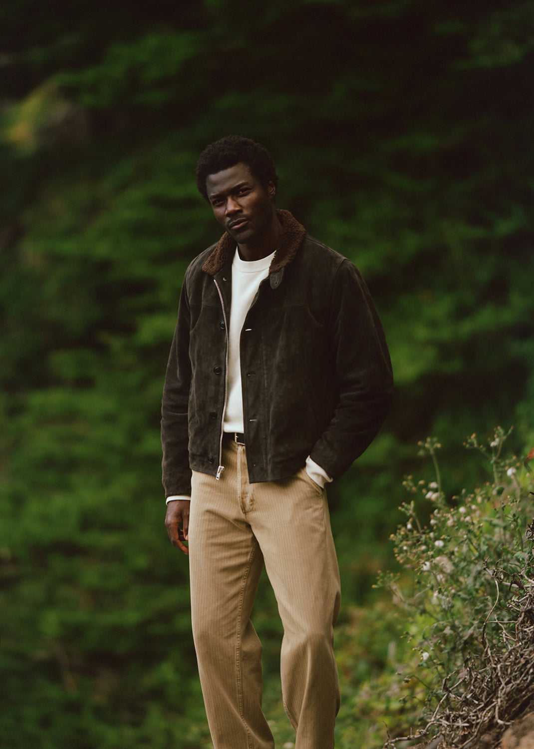 A man wearing a dark brown suede jacket with a brown shearling collar, a white t-shirt, and beige trousers, standing outdoors against a blurred green forest background.