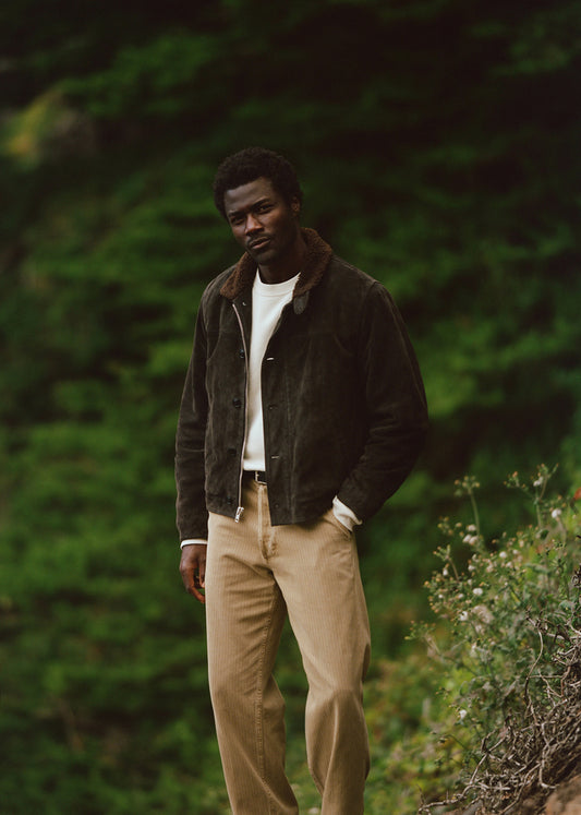 A man wearing a dark brown suede jacket with a brown shearling collar, a white t-shirt, and beige trousers, standing outdoors against a blurred green forest background.