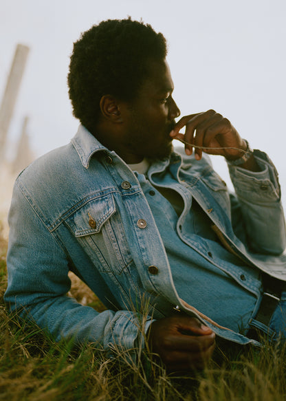 A man wearing a light blue denim jacket sits in grass outdoors.
