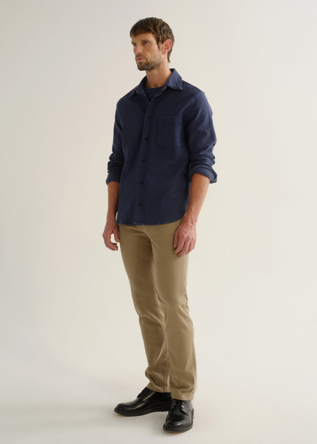 Person wearing a navy long sleeve button-up shirt with khaki pants and black shoes, angled front full body view on a light grey background.
