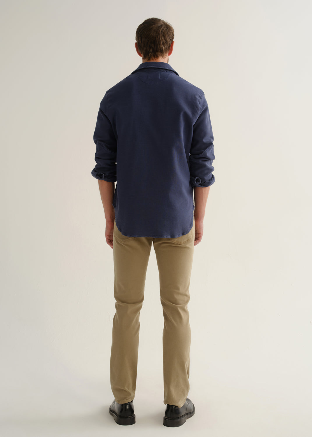 Person wearing a navy long sleeve button-up shirt with khaki pants and black shoes, back full body view on a light grey background.