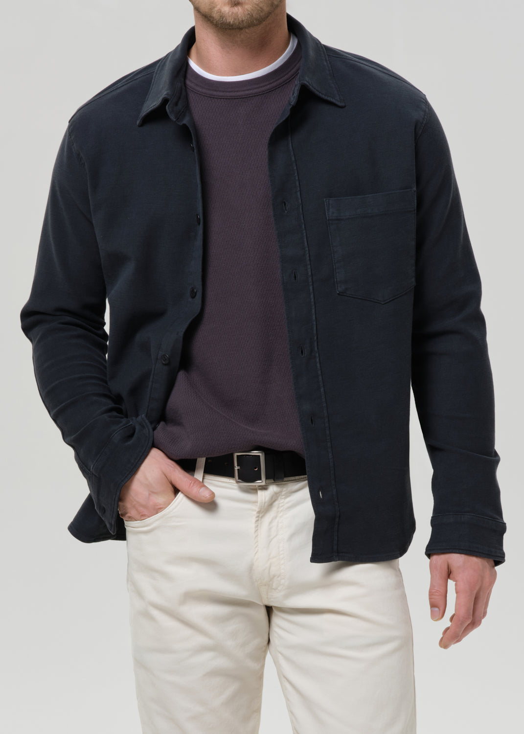 A man wearing a dark navy long-sleeve button-up overshirt over a dark plum crewneck sweater, paired with off-white trousers and a black belt, against a grey background.