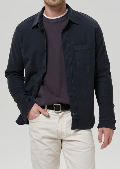 A man wearing a dark navy long-sleeve button-up overshirt over a dark plum crewneck sweater, paired with off-white trousers and a black belt, against a grey background.