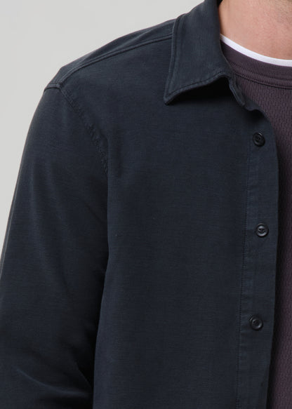 Close-up of a person wearing a dark navy button-up shirt over a dark ribbed knit top, against a grey background.