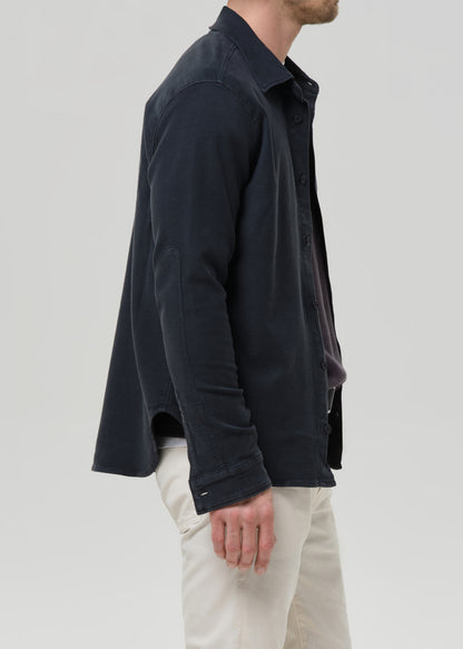 A man shown from the side wearing a dark navy long-sleeve button-up shirt and off-white trousers, photographed against a grey background.