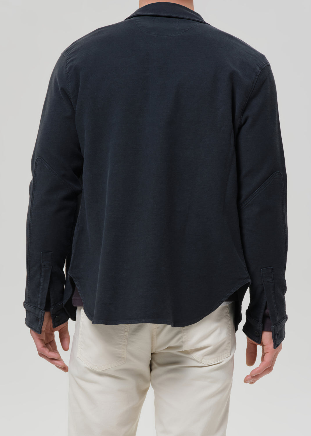 A man viewed from the back wearing a dark navy long-sleeve button-up shirt and off-white trousers, against a grey background.