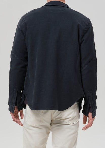 A man viewed from the back wearing a dark navy long-sleeve button-up shirt and off-white trousers, against a grey background.