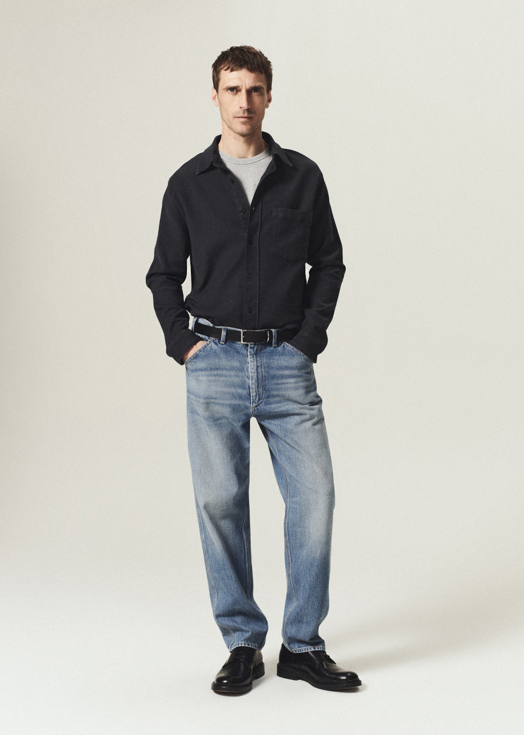 Man wearing a black button-up shirt layered over a grey crewneck top with light blue jeans on a grey background.