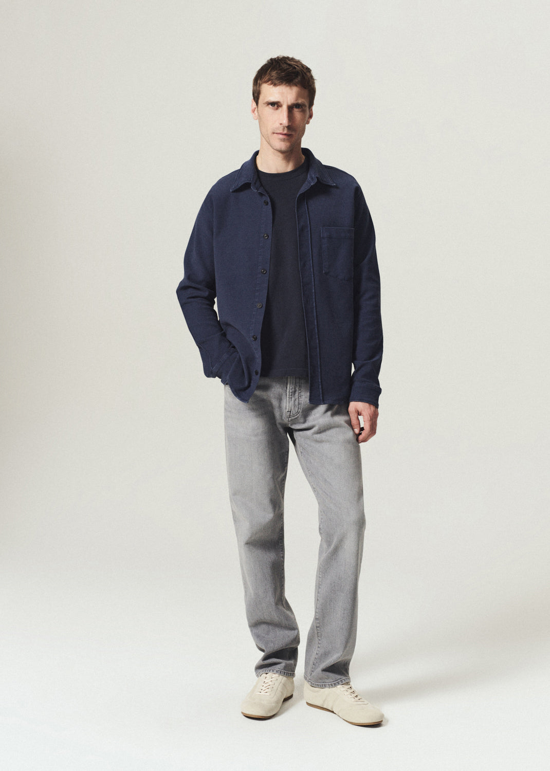Man wearing a navy button-up shirt layered over a navy crewneck top with grey jeans on a grey background.