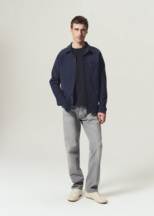 Man wearing a navy button-up shirt layered over a navy crewneck top with grey jeans on a grey background.