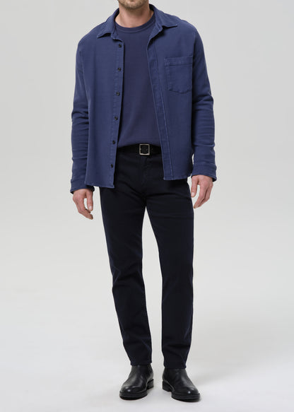 Person wearing a blue shirt over a dark shirt with black pants on a grey background