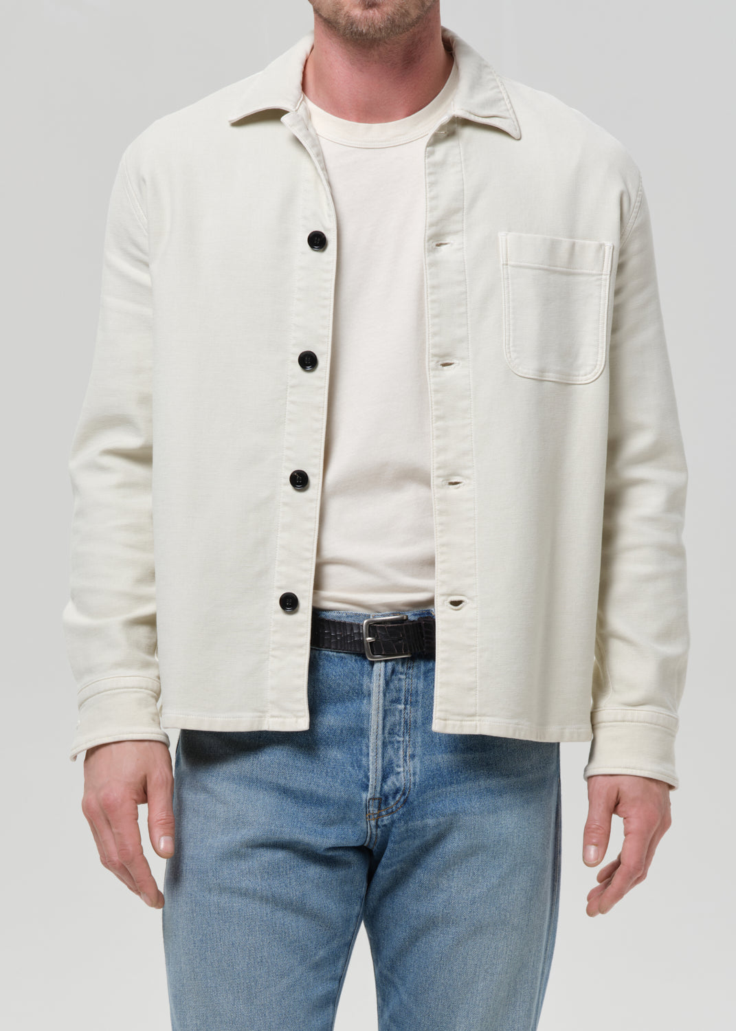 A man wearing a light cream-colored overshirt with black buttons over a matching cream t-shirt and blue jeans, photographed against a grey background.