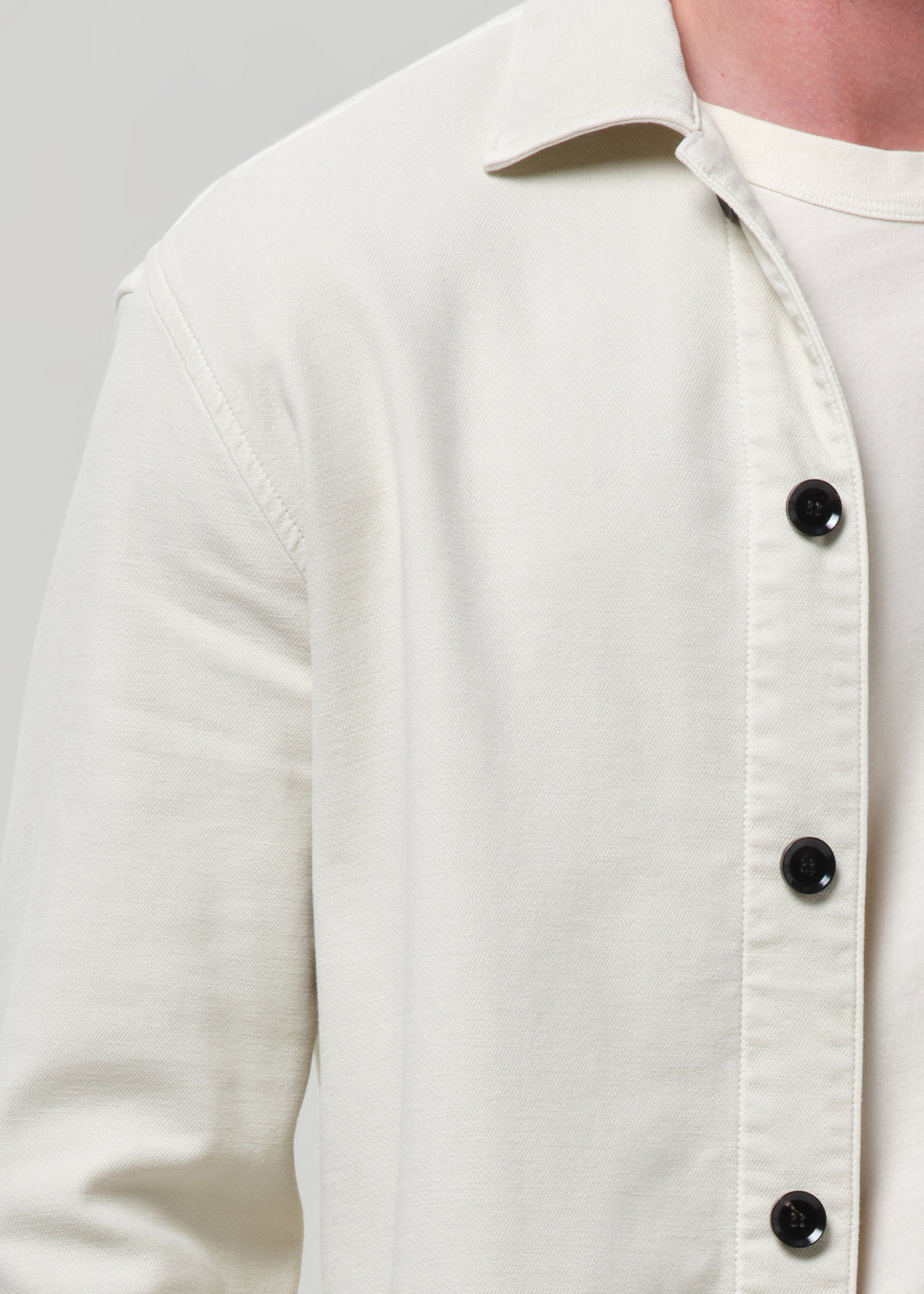 Close-up of a person wearing a cream-colored long-sleeve button-up shirt over a white undershirt, showing the collar, fabric texture, and black buttons against a grey background.