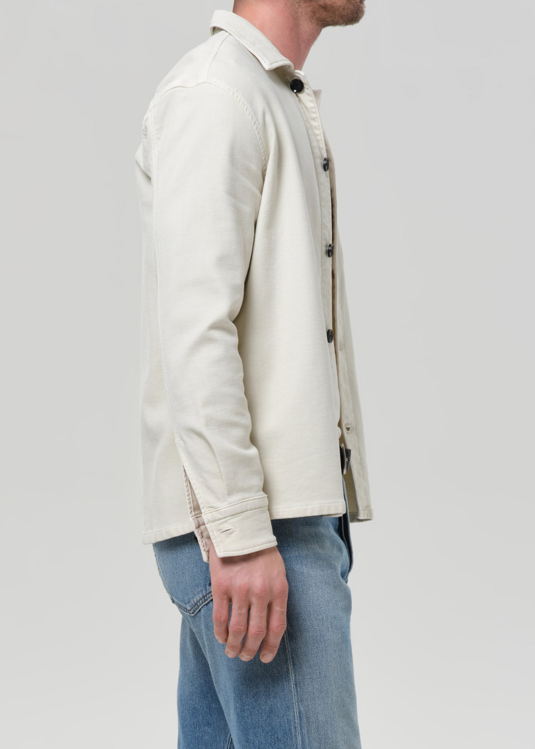 A man shown in side profile wearing a light cream-colored long-sleeve button-up shirt and blue jeans against a grey background.