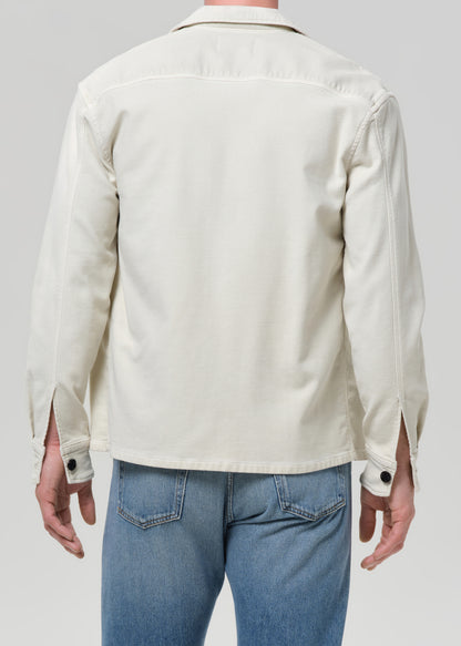 A man viewed from the back wearing a light cream-colored long-sleeve button-up shirt and blue jeans, set against a grey background.