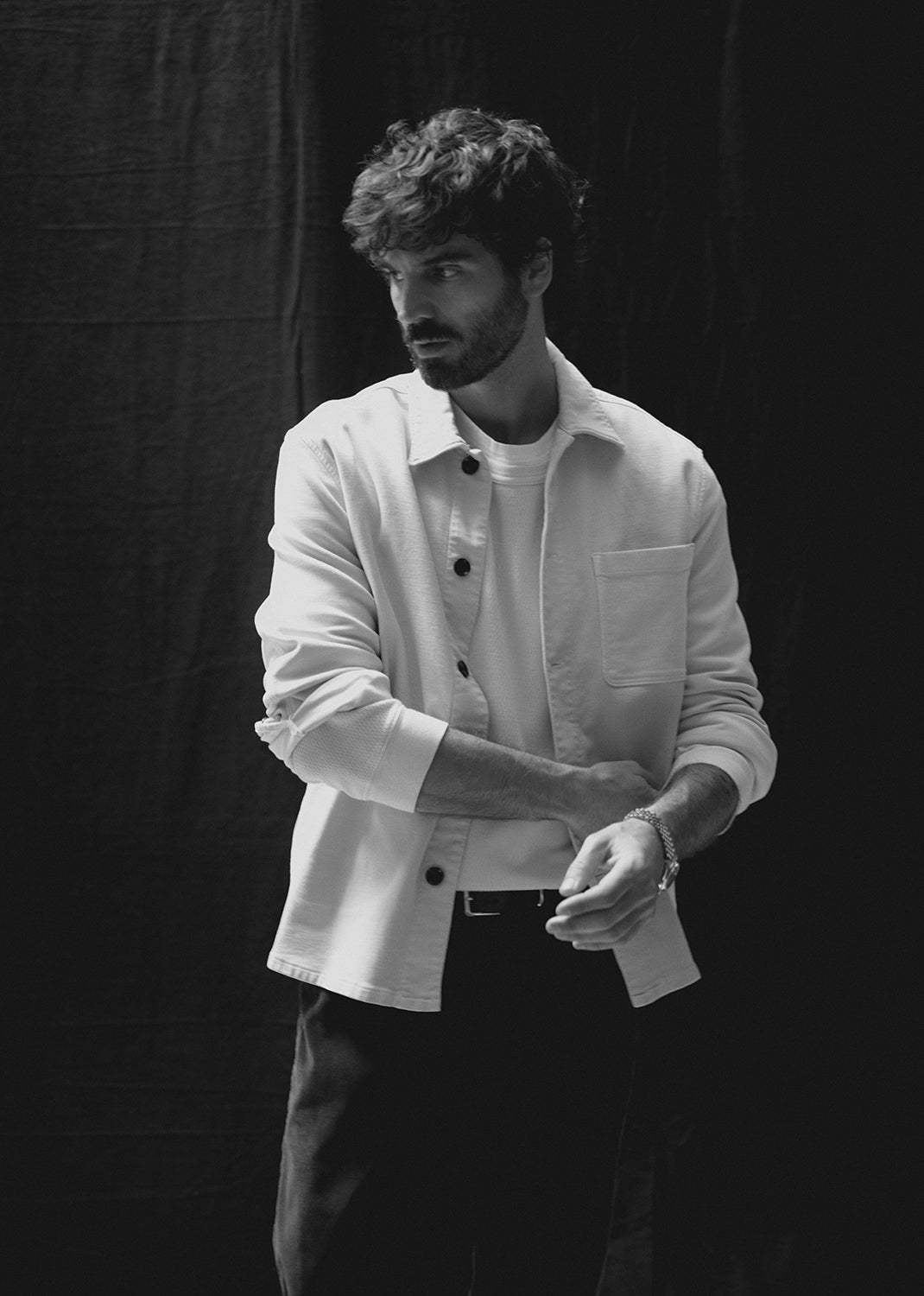 A man wearing a white button-up shirt over a white t-shirt and dark trousers, standing against a dark studio backdrop.