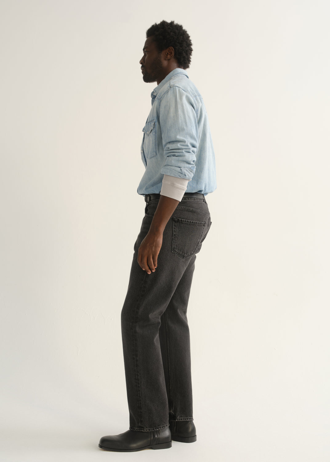 Person wearing a light blue denim button-front shirt, washed black jeans, and black boots, side full body view on a light grey background.