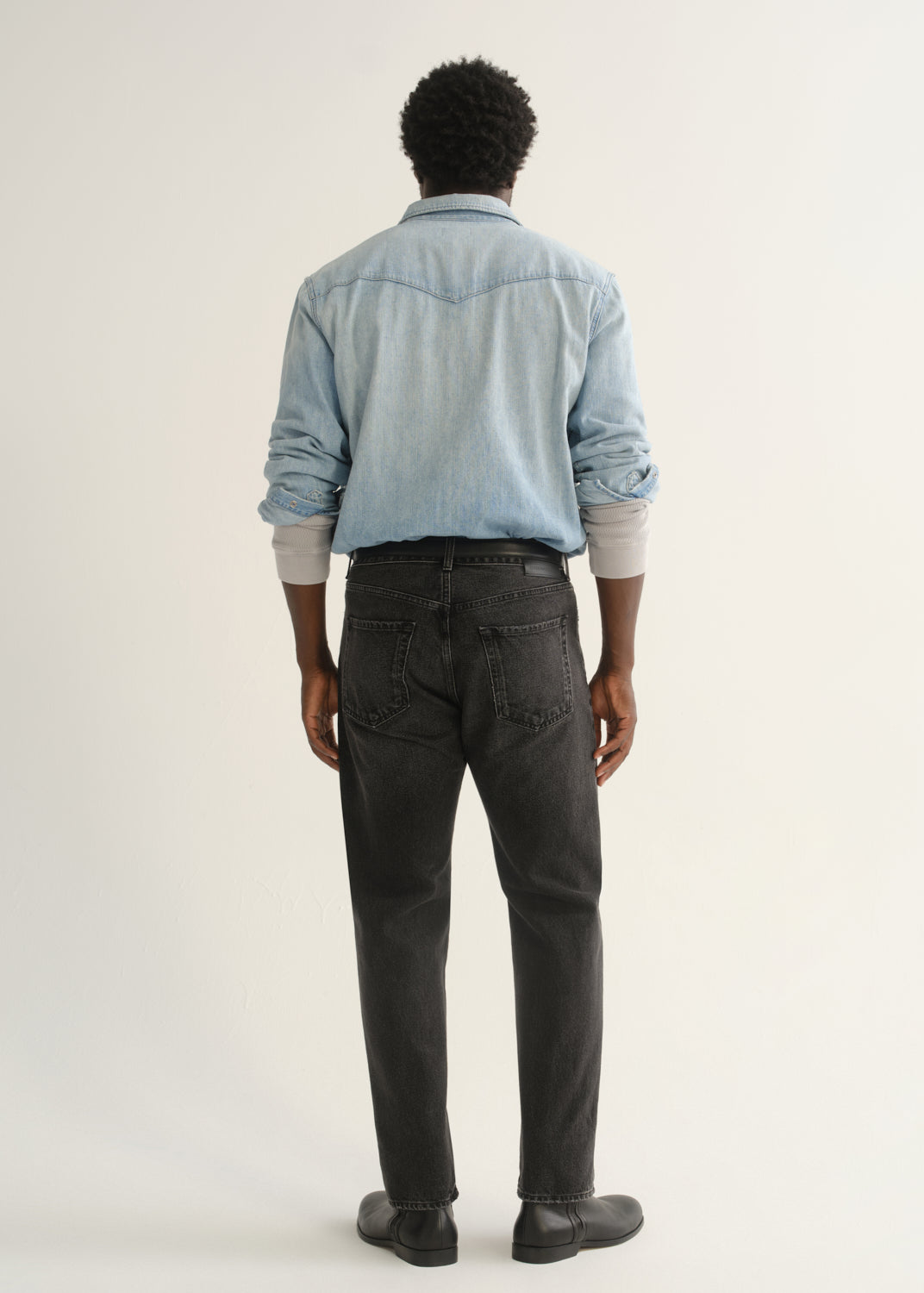 Person wearing a light blue denim button-front shirt, washed black jeans, and black boots, back full body view on a light grey background.