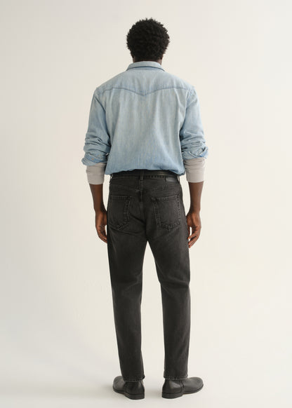 Person wearing a light blue denim button-front shirt, washed black jeans, and black boots, back full body view on a light grey background.