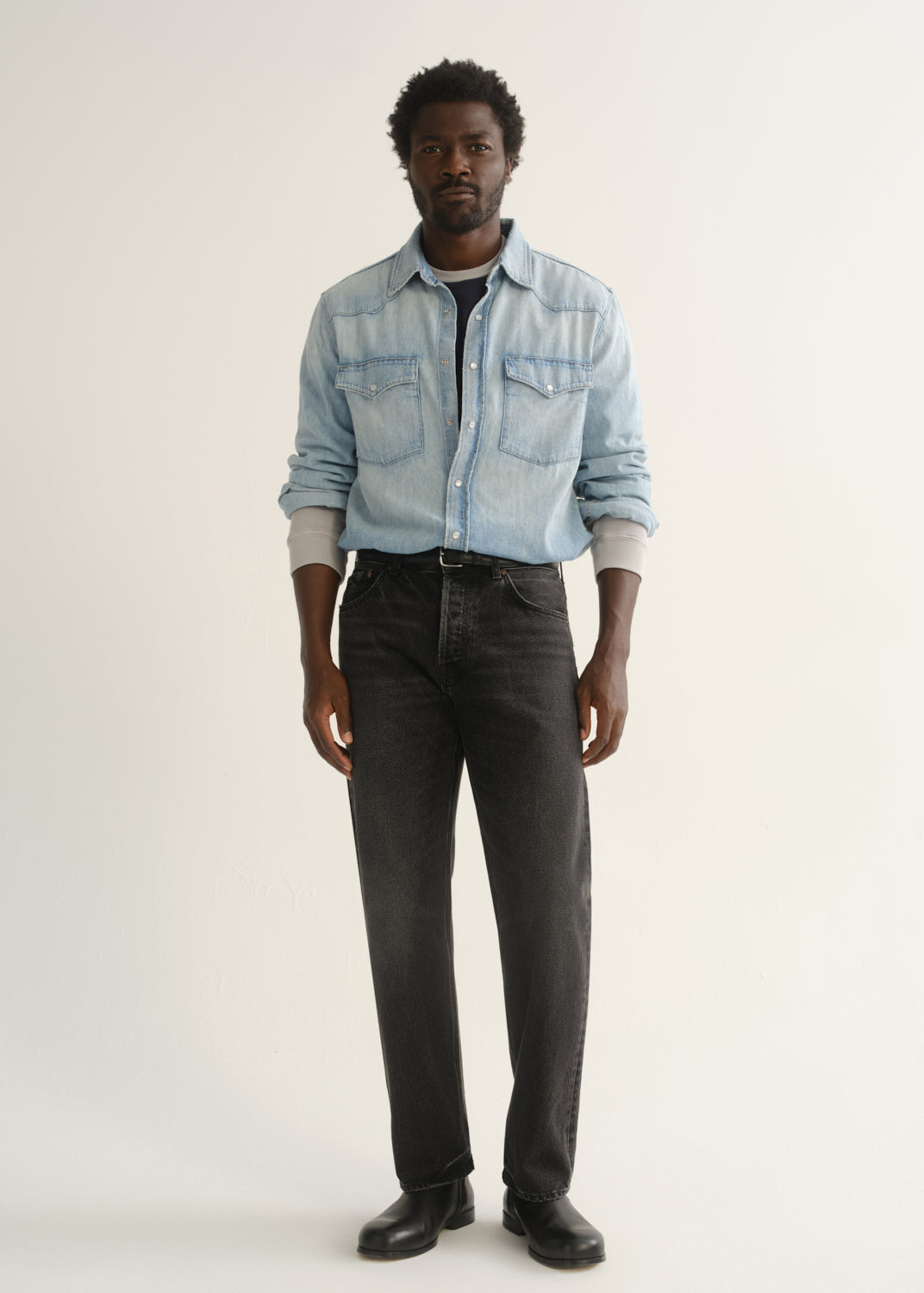 Person wearing a light blue denim button-front shirt, washed black jeans, and black boots, angled front full body view on a light grey background.