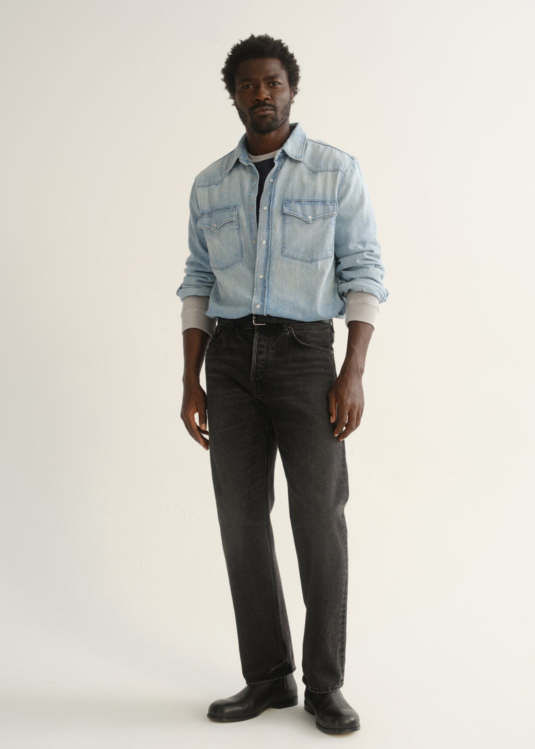 Person wearing a light blue denim button-front shirt, washed black jeans, and black boots, angled front full body view on a light grey background.