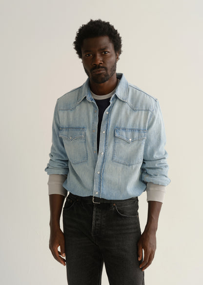 Person wearing a light blue denim button-front shirt, washed black jeans, and black boots, front full body view on a light grey background.