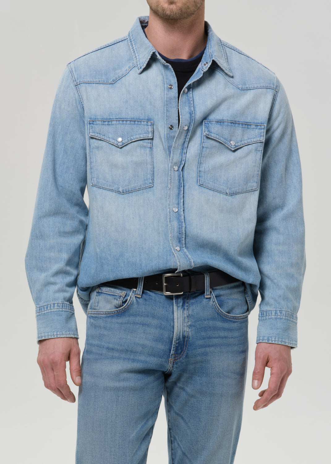 A man wearing a light blue denim western-style shirt jacket with two chest pockets, paired with blue jeans and a black belt, shown from the neck down against a grey background.
