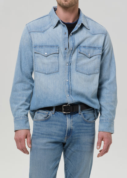 A man wearing a light blue denim western-style shirt jacket with two chest pockets, paired with blue jeans and a black belt, shown from the neck down against a grey background.