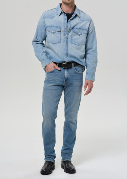 A man wearing a light blue denim western shirt and matching light blue jeans, styled with a black belt and black leather shoes, photographed against a grey background.