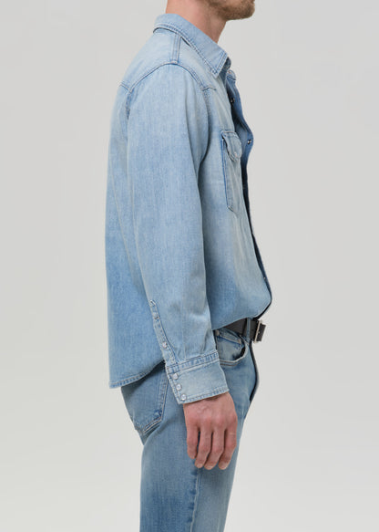 A side-profile view of a man wearing a light blue denim shirt jacket over a dark shirt, paired with matching light blue denim jeans, against a grey background.