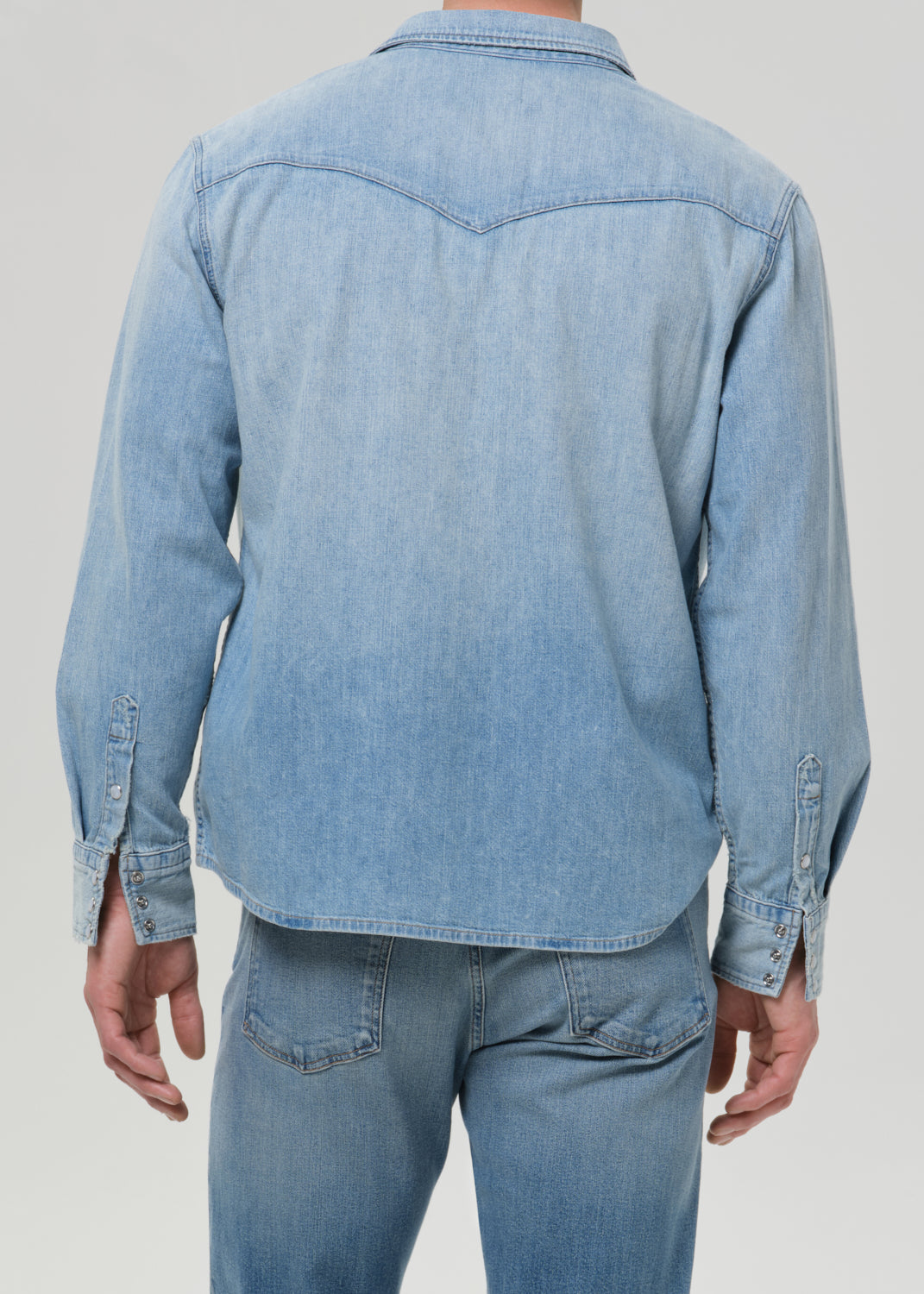 A man viewed from the back wearing a light blue denim long-sleeve button-up shirt and matching light blue denim jeans, against a grey background.