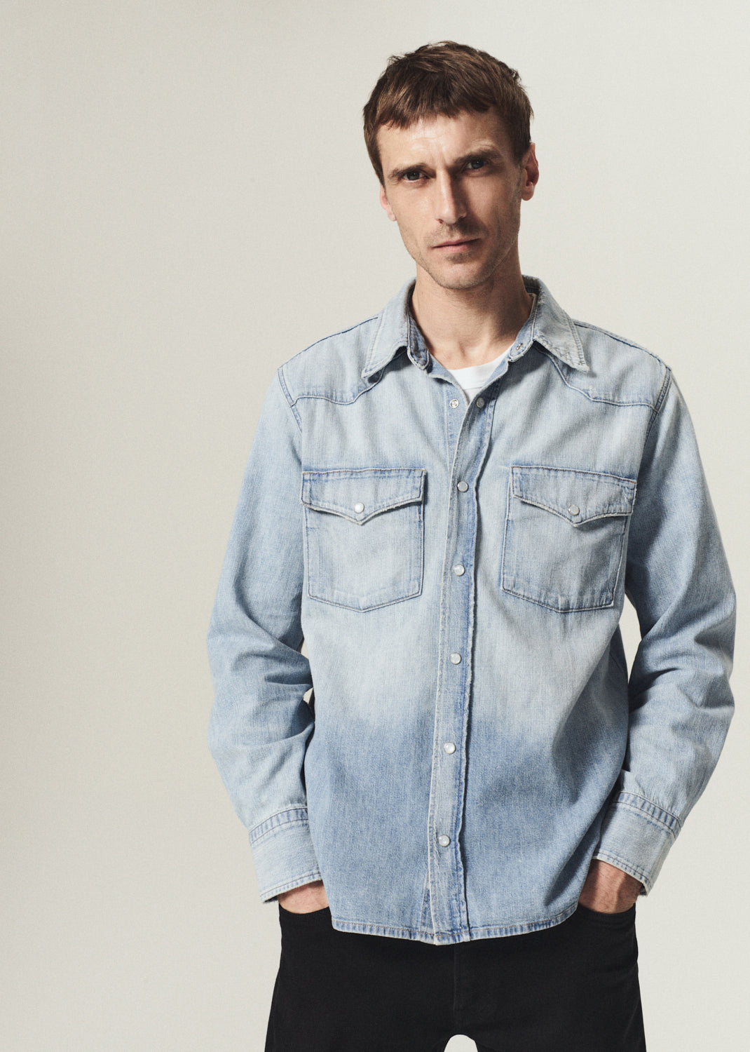 A man wearing a light blue denim western-style button-up shirt with two chest pockets, photographed against a plain light gray studio background.