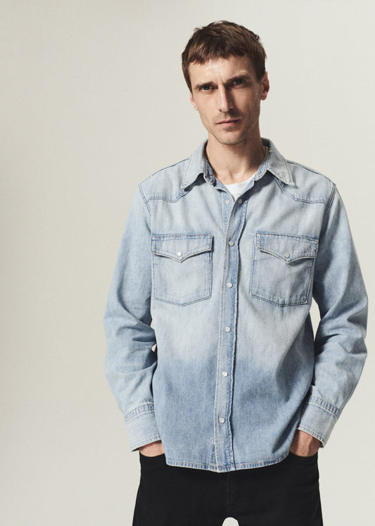 A man wearing a light blue denim western-style button-up shirt with two chest pockets, photographed against a plain light gray studio background.