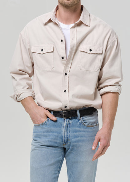 A man wearing a light beige long-sleeve button-up shirt with two chest pockets, tucked into light blue jeans with a black belt, against a grey background.