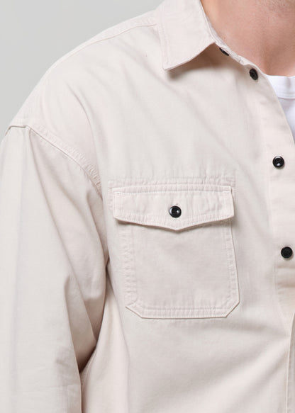 Close-up of a person wearing a light beige or cream-colored long-sleeve button-up shirt with a chest pocket and black buttons, layered over a white undershirt.