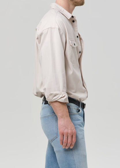 A man shown in side profile wearing a light beige long-sleeve button-up shirt with rolled-up sleeves and light blue jeans, against a grey background.