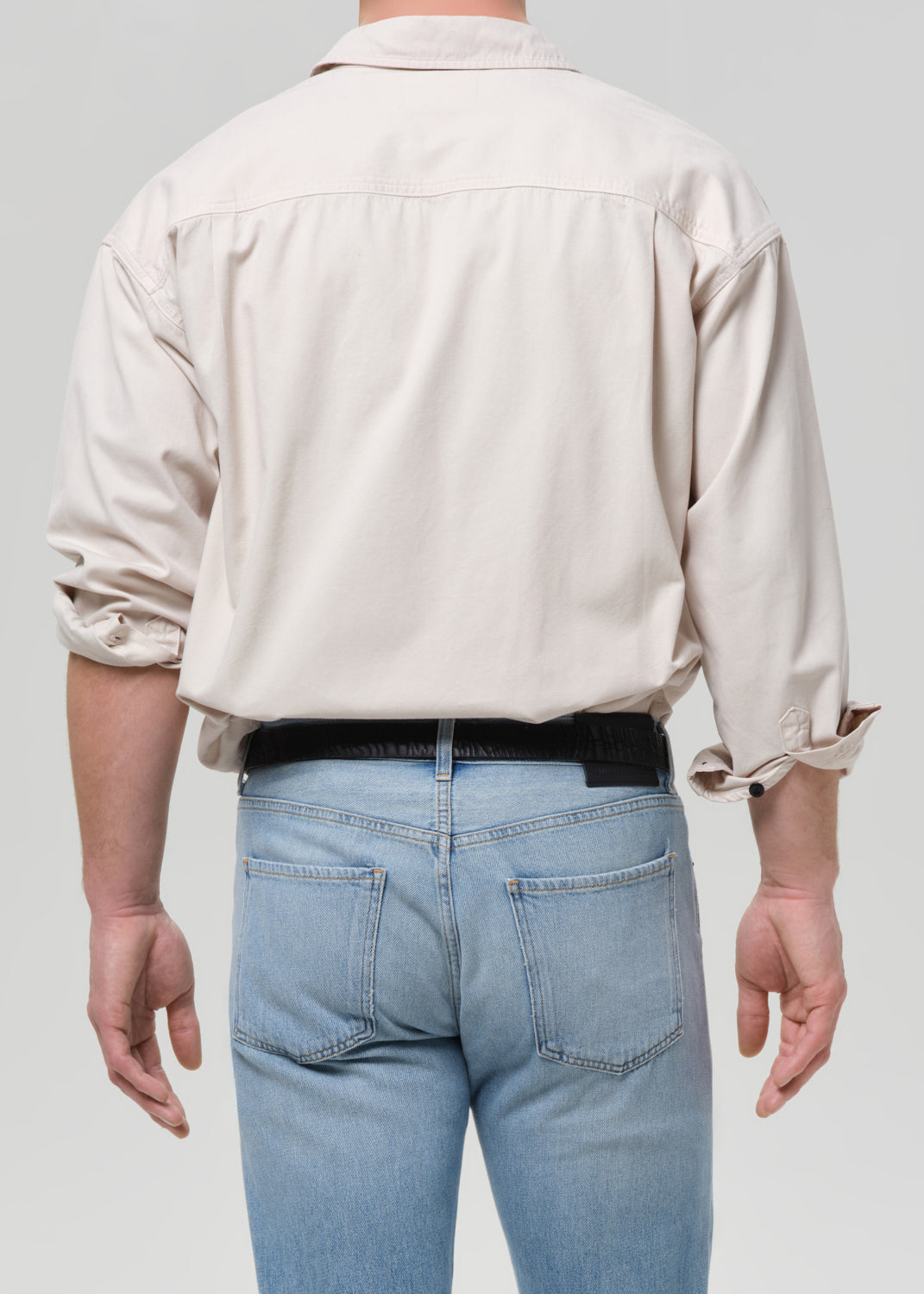 Rear view of a person wearing a light beige long-sleeve button-up shirt and light blue jeans with a black belt, against a grey background.