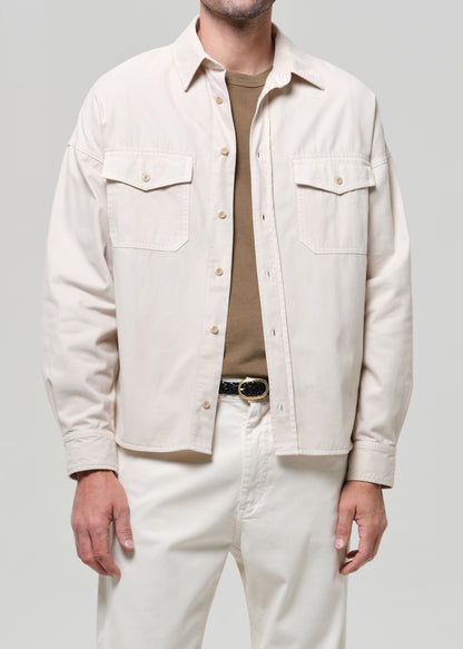 Man wearing a beige shirt over a brown shirt with white pants on a plain background