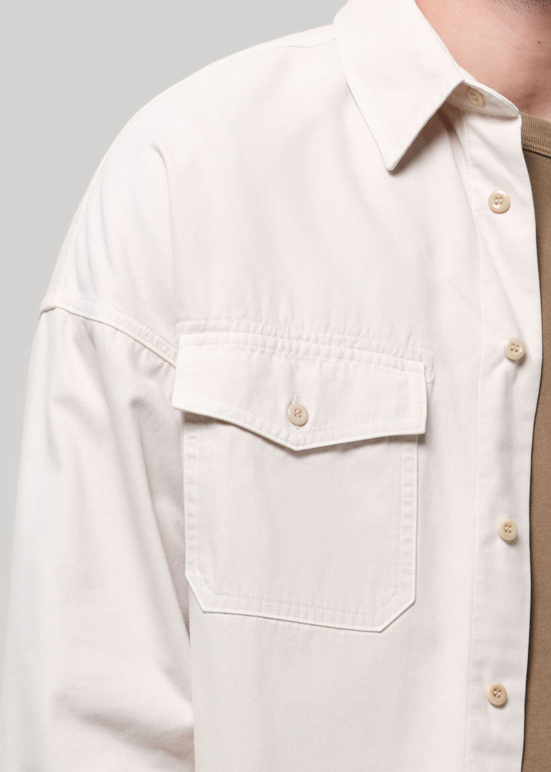 Man wearing a beige shirt over a brown shirt with white pants on a plain background