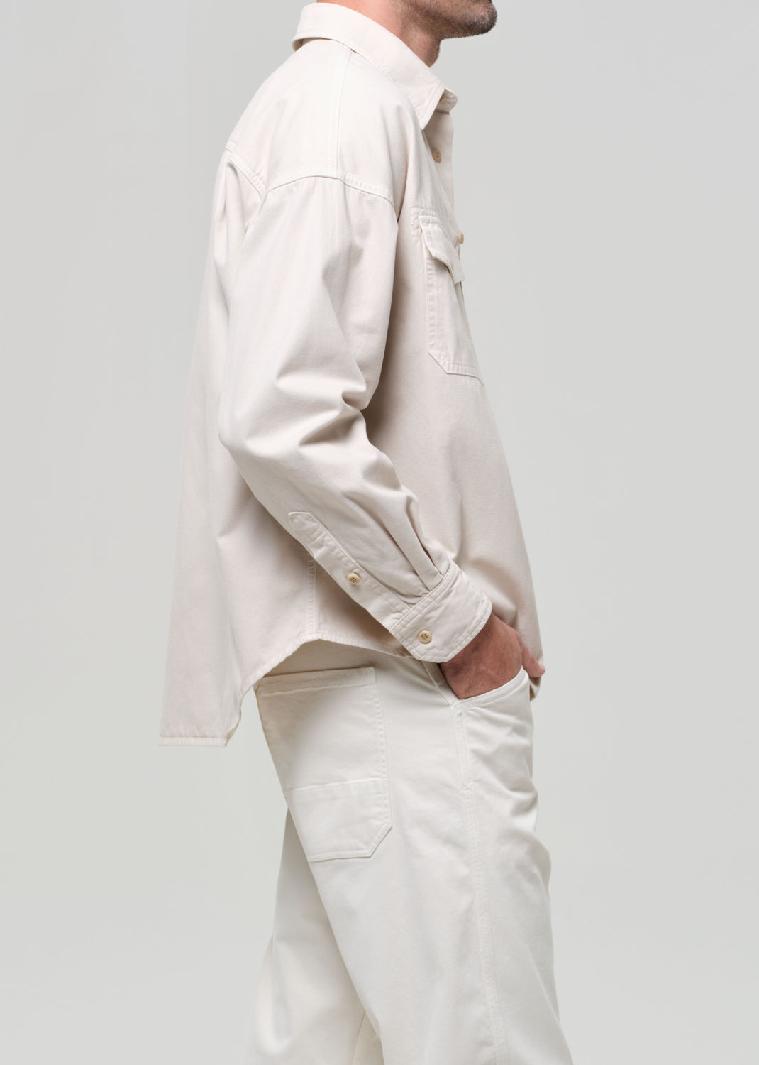 Man wearing a beige shirt over a brown shirt with white pants on a plain background
