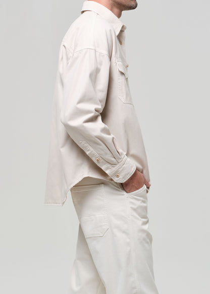 Man wearing a beige shirt over a brown shirt with white pants on a plain background
