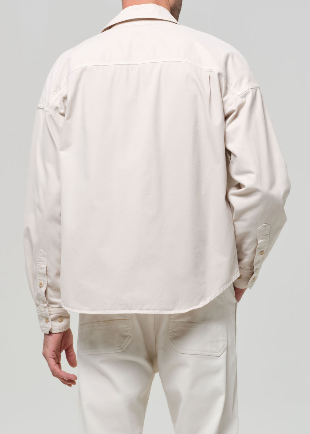 Man wearing a beige shirt over a brown shirt with white pants on a plain background