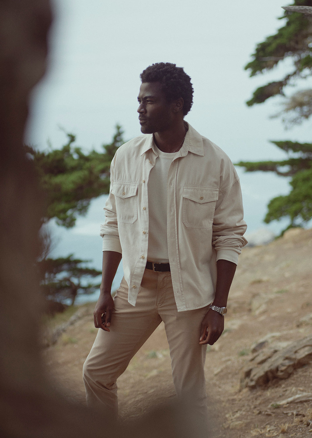 A man wearing a light beige button-up shirt over a white t-shirt and beige pants, standing outdoors on a rocky hillside with trees in the background.
