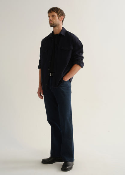 Person wearing a dark navy long sleeve button-up shirt with dark blue jeans and black boots, angled front full body view on a light grey background.