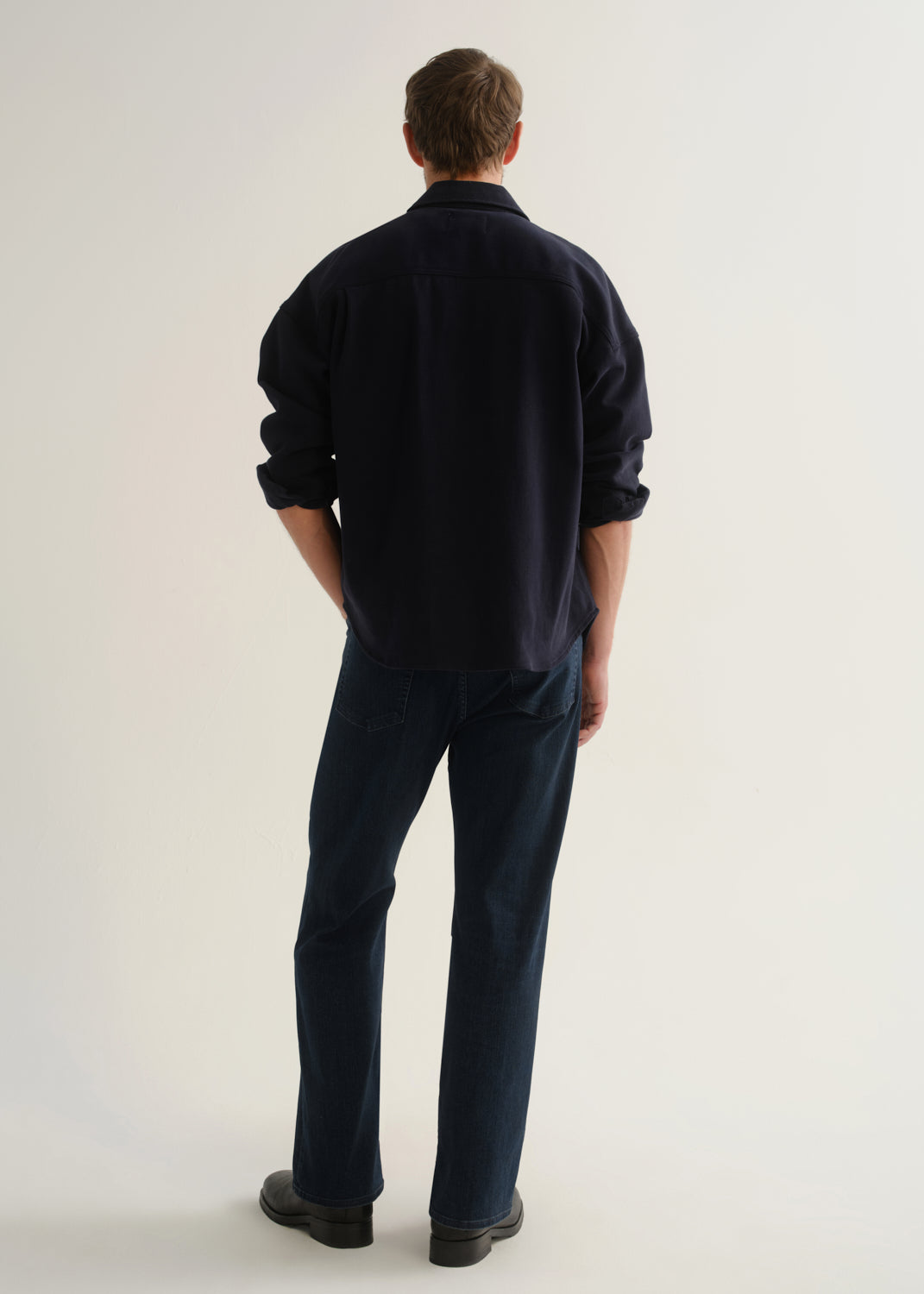 Person wearing a dark navy long sleeve button-up shirt with dark blue jeans and black boots, back full body view on a light grey background.